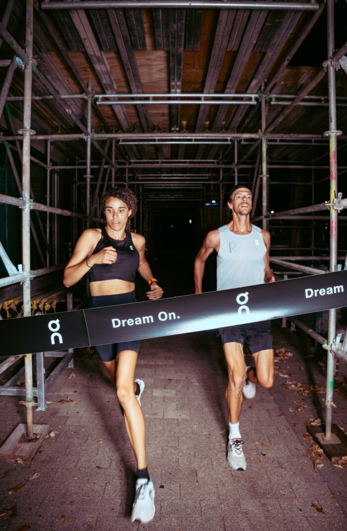 Two runners running through scaffolding about to break finish line tape.