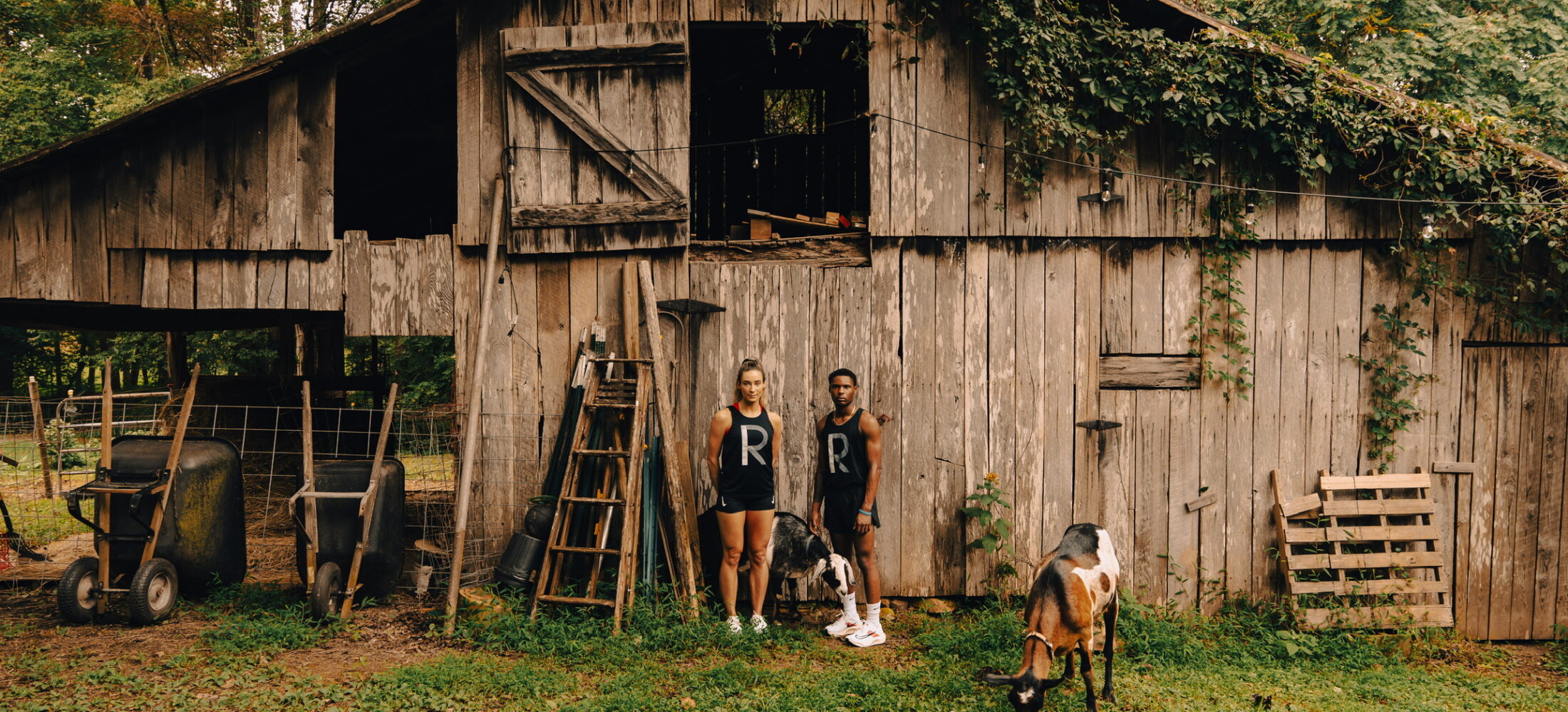 Two runners and a goat standing in front of a run down barn